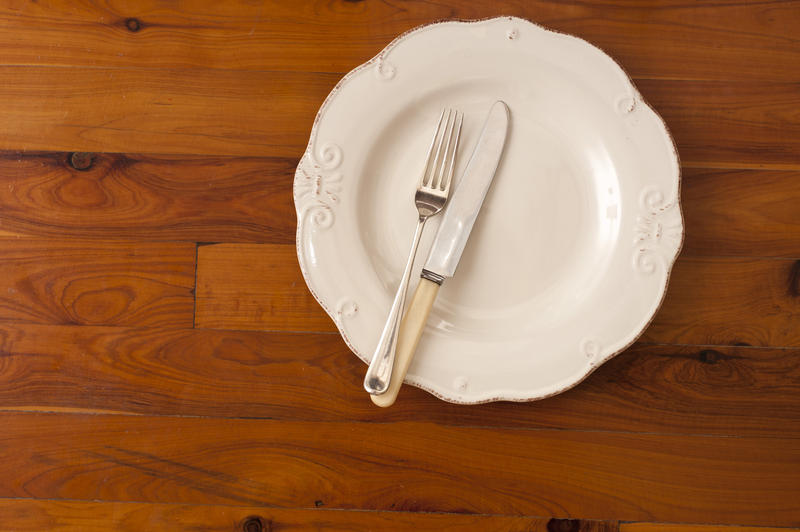 Overhead view of a plain white empty dinner plate with a knife and fork on it standing on a wooden dining table with copyspace