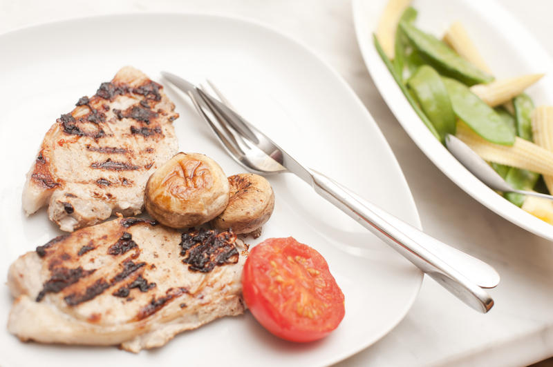 Grilled pork chops served with tomato and mushrooms and a side plate of and vegetables containging mangetout peas and baby corn