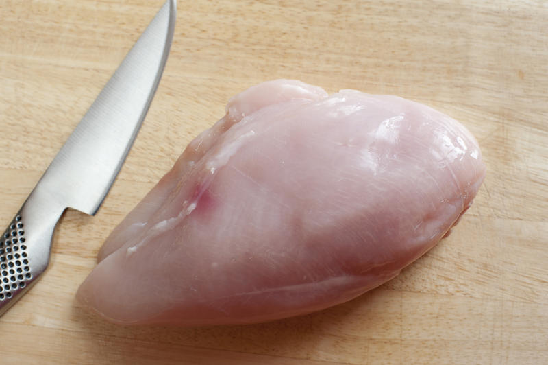 Preparing raw chicken breast for cooking with an uncooked breast lying on a wooden kitchen counter with a sharp stainless steel knife