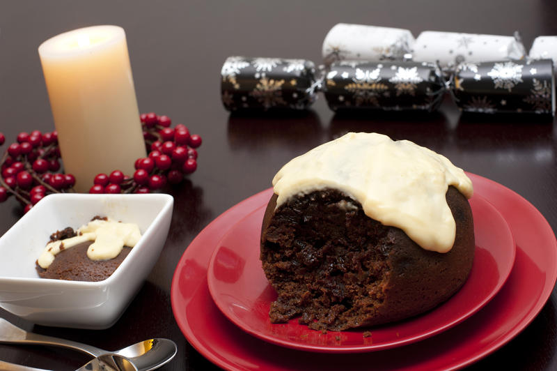 Serving of traditional steamed fruity Christmas pudding and brandy custard on a festive table with a glowing seasonal candle and wreath of colourful red berries