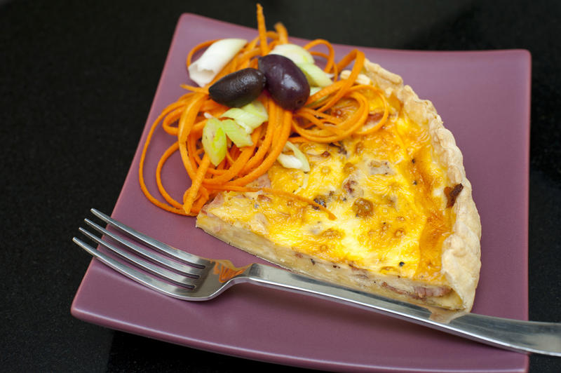 Slice of savoury quiche baked with an egg custard base and served with a side portion of spiced spaghetti and olives