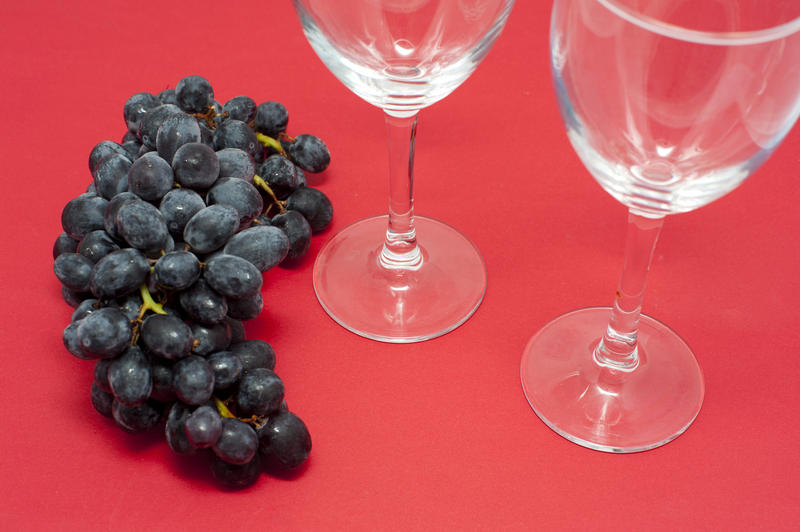 Close Up of Bunch of Dark Red Grapes on Red Studio Background Next to Two Empty Wine Glasses with Copy Space