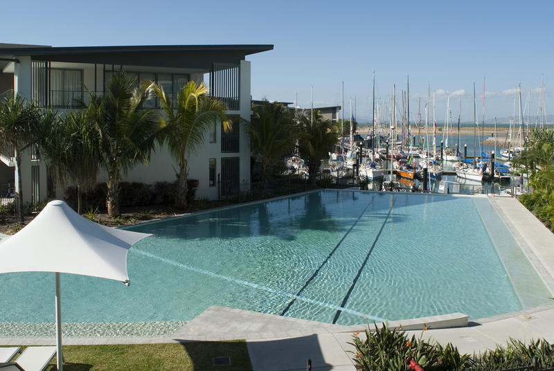 Sparkling blue swimming pool and umbrellas at a tropical resort with yachts moored in a marina or harbour in the background