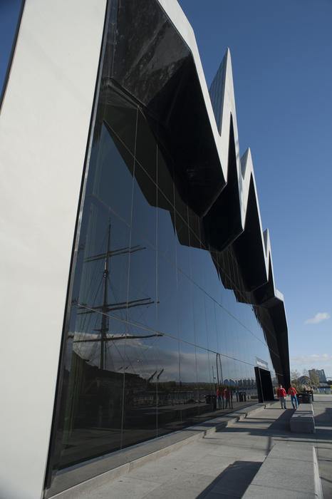 External facade of the Glasgow Riverside Museum with its distinctive modern architecture and a tall ship in the harbour reflected in the window