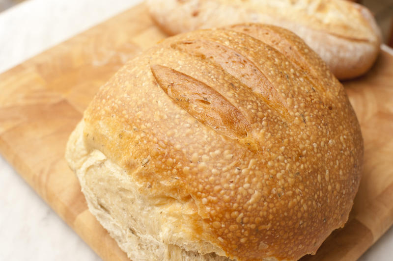 Round loaf of fresh rustic bread with a crusty top on a wooden bread board, close up view from above