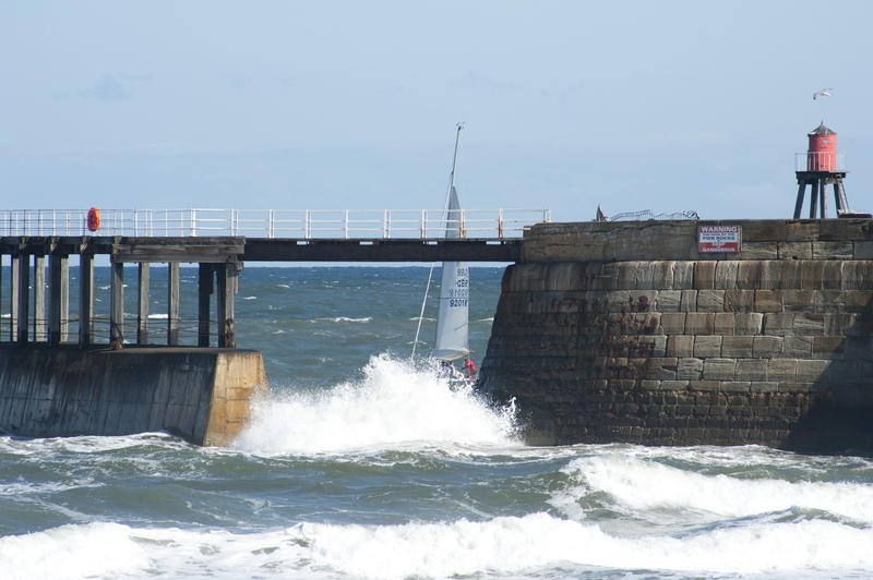 Sailing yacht passing through the sheltered waters behind the Whitby breakwater and stone pier with turbulent surf in the foreground
