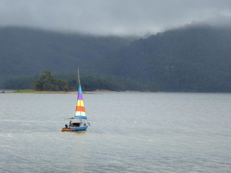 Small sailboat with a single colorful sail cruising offshore in a calm ocean on a misty day with mountains visible in the distance