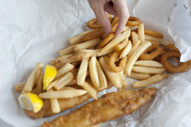 Hand of a man eating an unhealthy deep fried fish and chips takeaway meal off a sheet of crumpled white paper