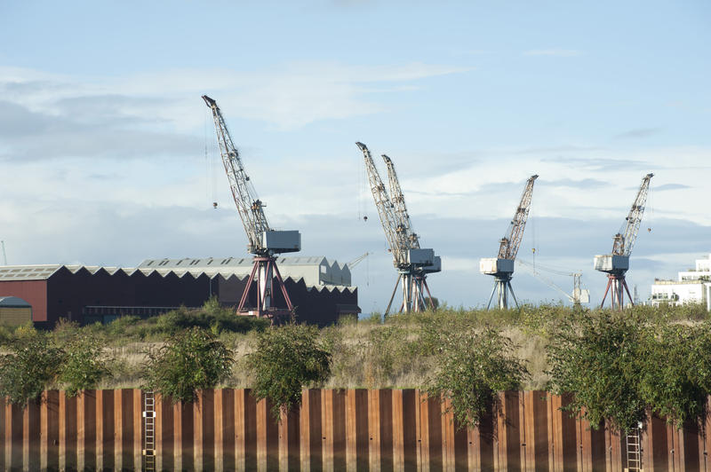 Large industrial dockyard cranes and warehouses at a shipbuilding yard viewed over a metal perimeter fence