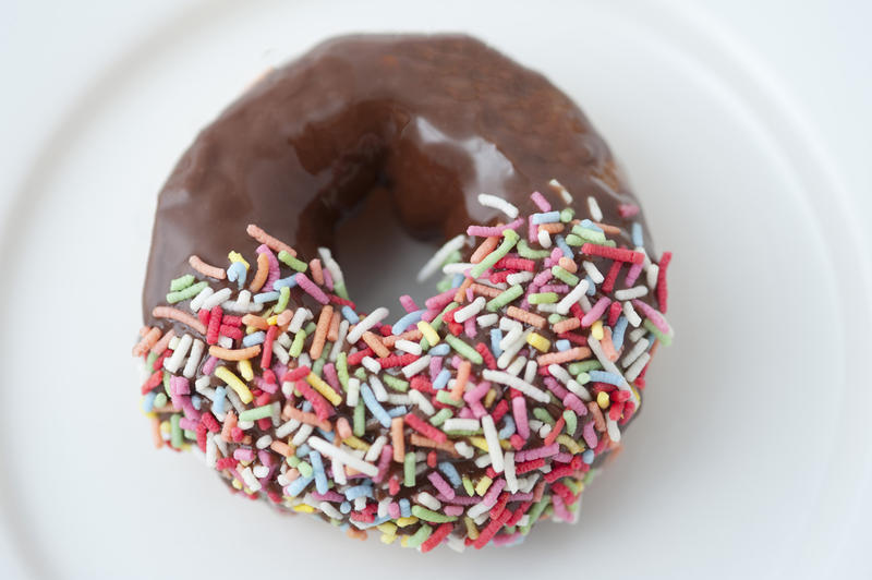Freshly baked single chocolate doughnut dipped in multicolored candy sprinkles served on a white plate for a morning coffee break