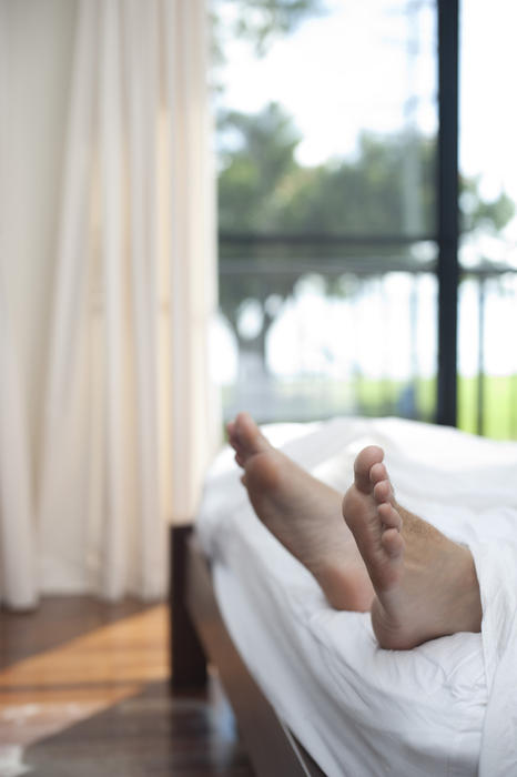 Man lying in having a lazy day in bed with his bare feet sticking out from the end of the white bedclothes, close up view against a view window