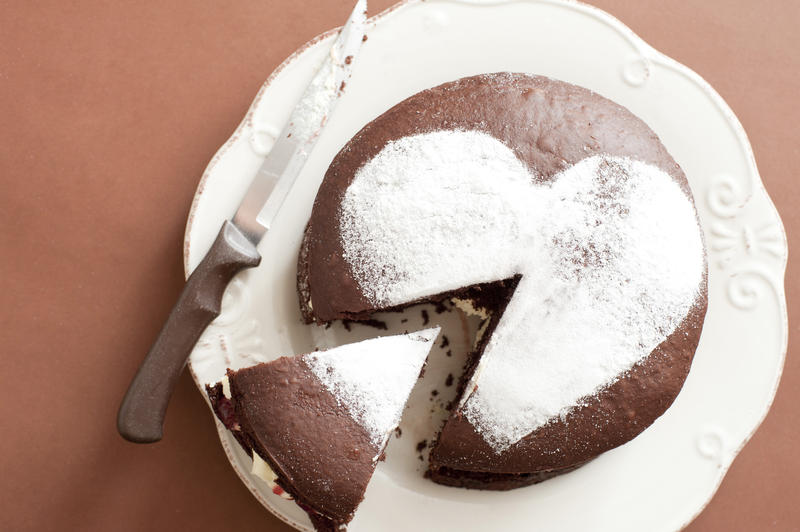 Aerial Shot of Sliced Chocolate Cake with Heart Shape Sugar on Top, Served on White Plate with Knife on Side. Placed on brown Table.
