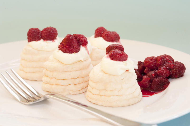 Individual small meringue pavlovas filled with whipped cream and topped with raspberries with a portion of raspberry compote on the side
