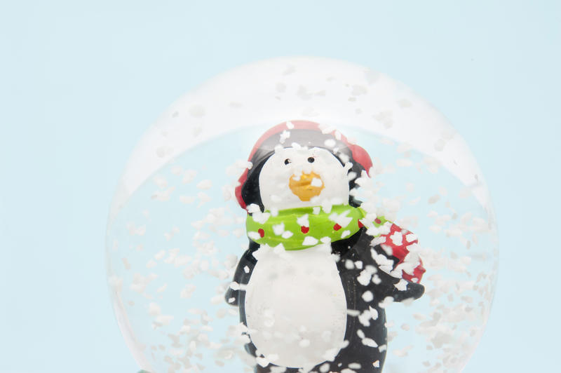 Cute little penguin in a scarf and winter cap standing in snowflakes floating in a spherical Christmas snow globe decoration
