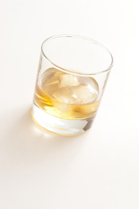Plain glass tumbler containing whiskey on the rocks at a tilted angle on a white background