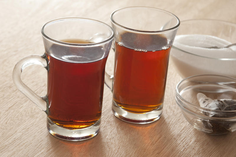Two clear glass mugs of hot sweet black tea with the used teabags in a dish alongside and a bowl of sugar behind