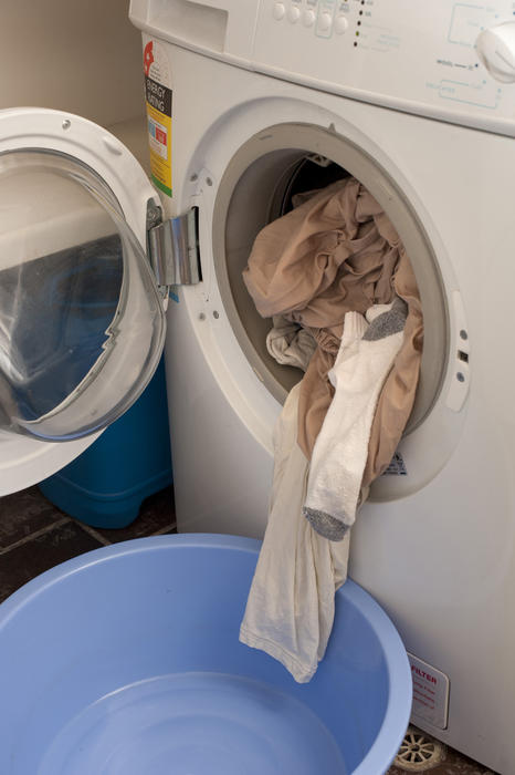 Clean washing spilling out of the open door of a front loader a washing machine into a blue plastic tub on the floor in a hygiene and household chores concept