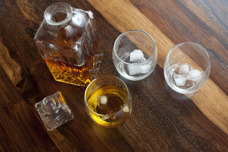 Whiskey on the rocks with an overhead view of a square decanter alongside a tumbler with whiskey and ice and two further glasses with ice. high angle view on a wooden bar counter