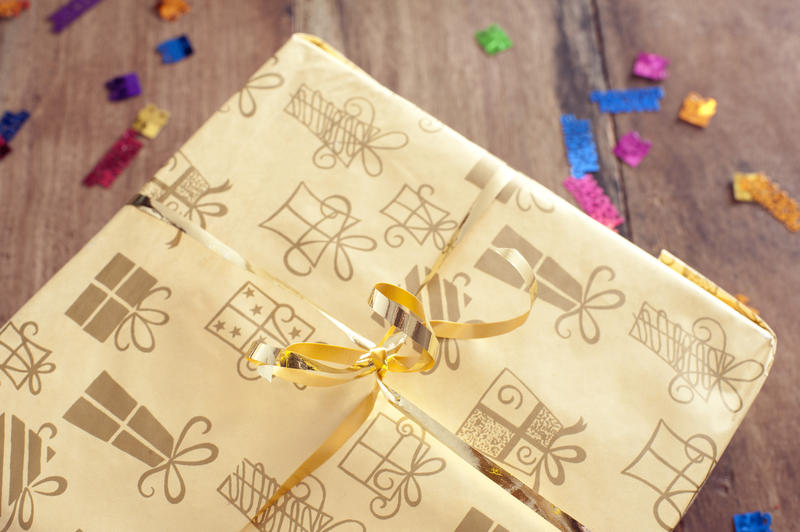 Wrapped gift with decorative patterned paper and a gold ribbon and bow standing on a wooden table with party confetti, close up view from the top