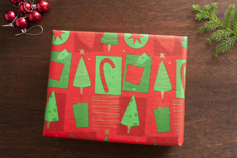 Colorful gift-wrapped Christmas present with red and green patterned paper lying on a wooden table alongside red baubles and a fresh green fir branch, overhead view
