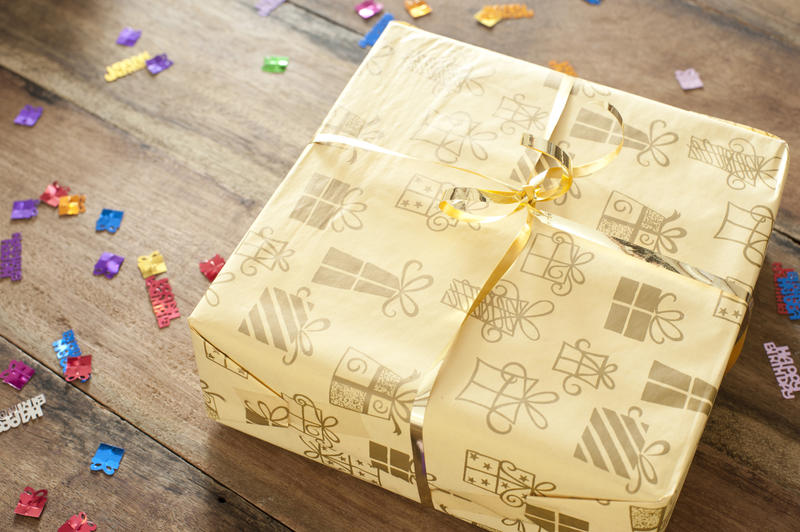 Close up Yellow Present Tied with Gold Ribbon on Top of a Wooden Table with Confetti Scattered.
