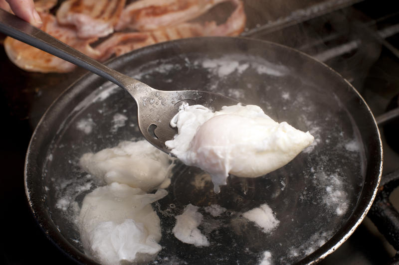 Cook removing a poached egg from the boiling water in a pan ready to serve for breakfast with bacon visible behind