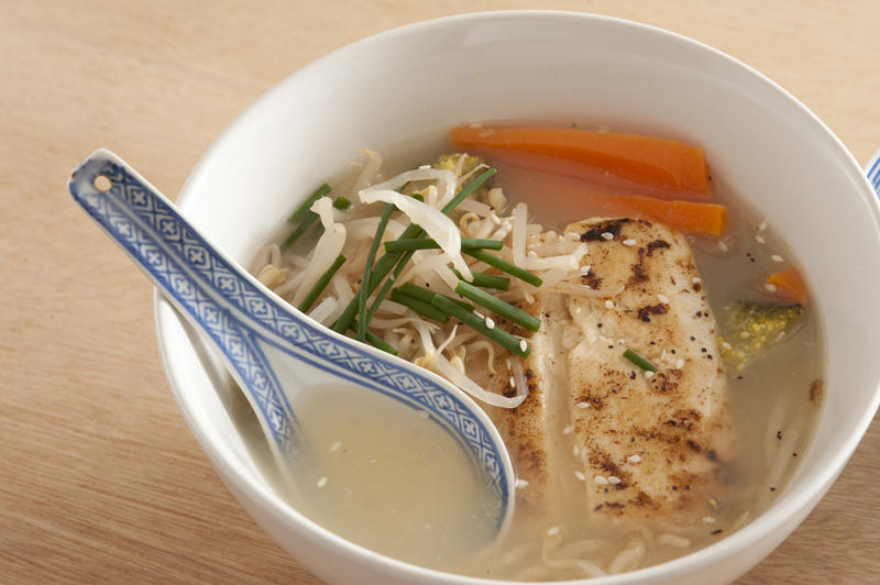 Close up on small white bowl of warm baked fish and noodle soup with garlic, chives, carrots and other herbs on wooden table