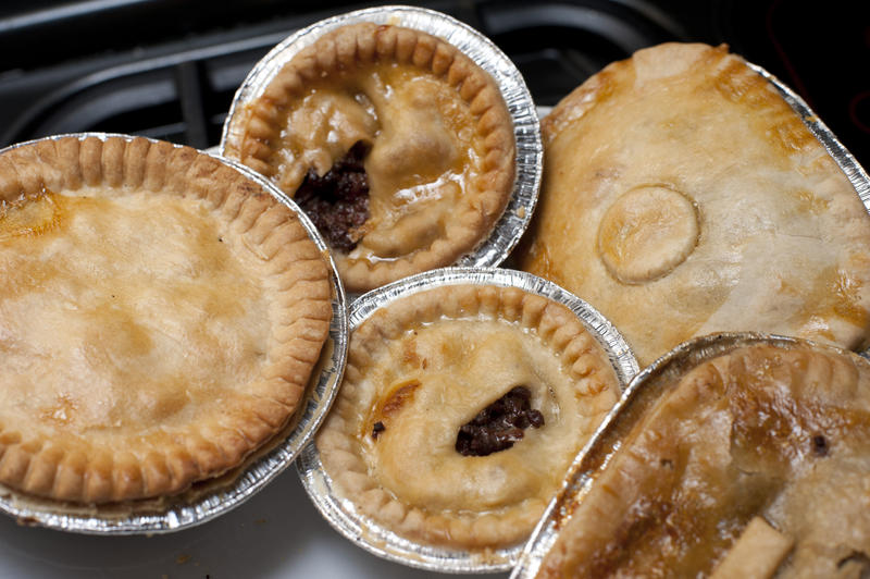 Assorted pastry meat pies in tin foil bases in a variety of shapes on a hot plate or oven tray ready for heating for lunch