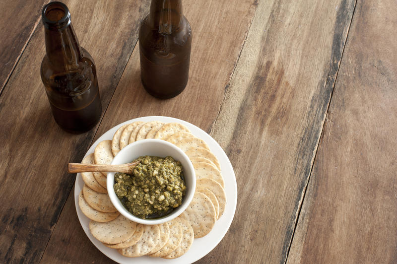 Delicious serving of green basil pesto with spoon and surrounded by water crackers beside two beer bottles