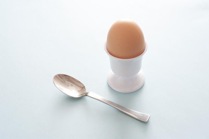 Boiled egg for breakfast in a plain white egg cup served with a silver spoon on light grey with copy space