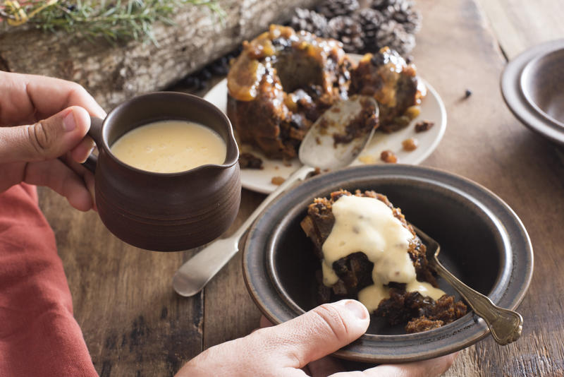 Man pouring brandy sauce from a small jug onto a helping of Christmas pudding in a bowl over the table