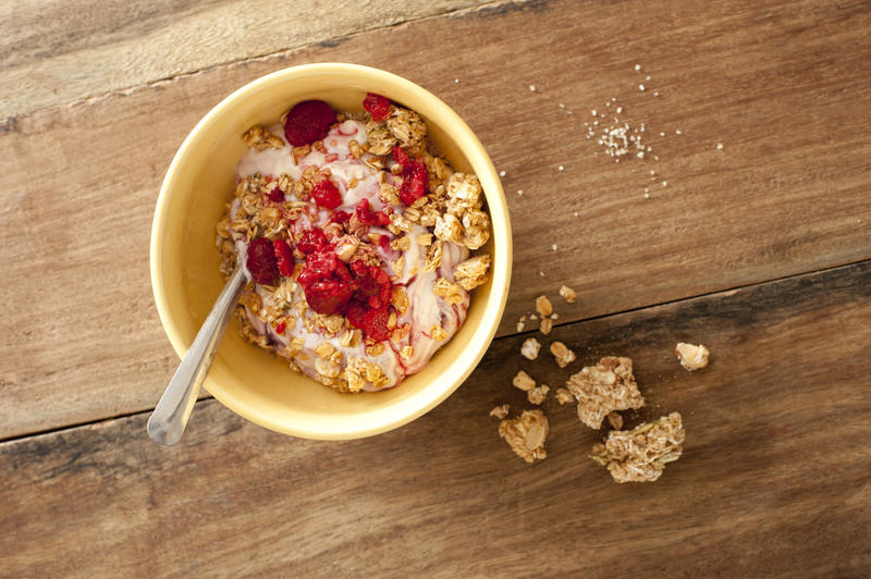 First person perspective view of delicious breakfast muesli in little yellow bowl with fruit and milk yogurt on table with some spilled on side