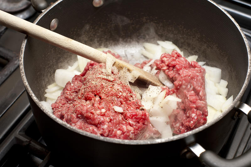 Browning beef mince with diced onions in a frying pan over a gas hob stirring with a wooden spoon