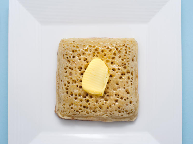 Overhead view of white square bowl with a golden crumpet and a dab of butter on top
