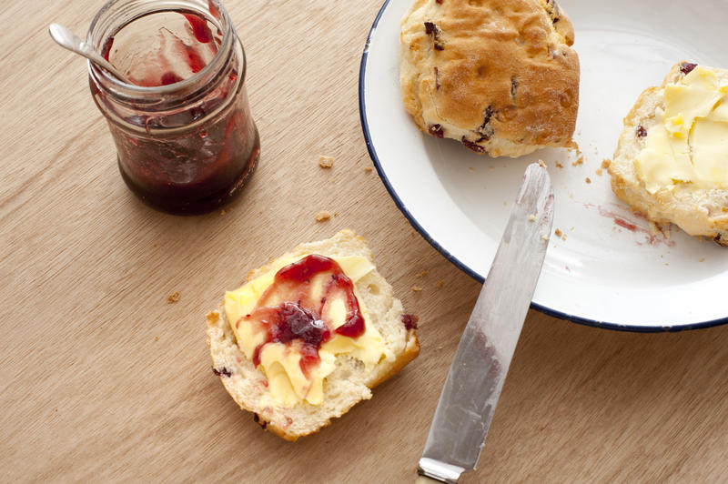 Buttered scone with strawberry jam on a wooden table alongside a jar of preserve with a plate and scones nearby