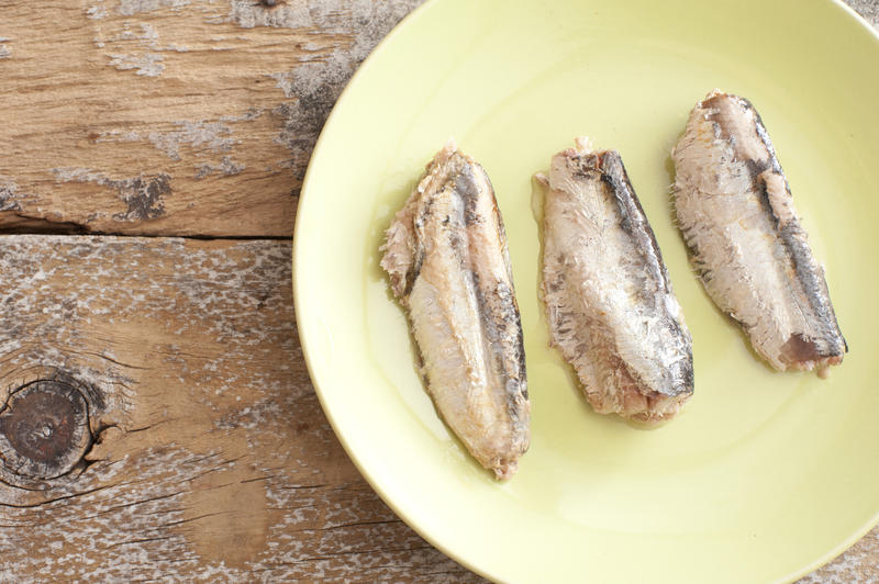 Three freshly unpacked oily sardines with bones on green plate over weathered wooden table