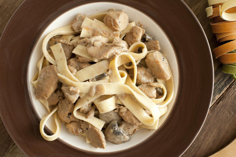 First person view on brown and white ceramic bowl of cooked ribbon pasta noodles with chicken meat on table