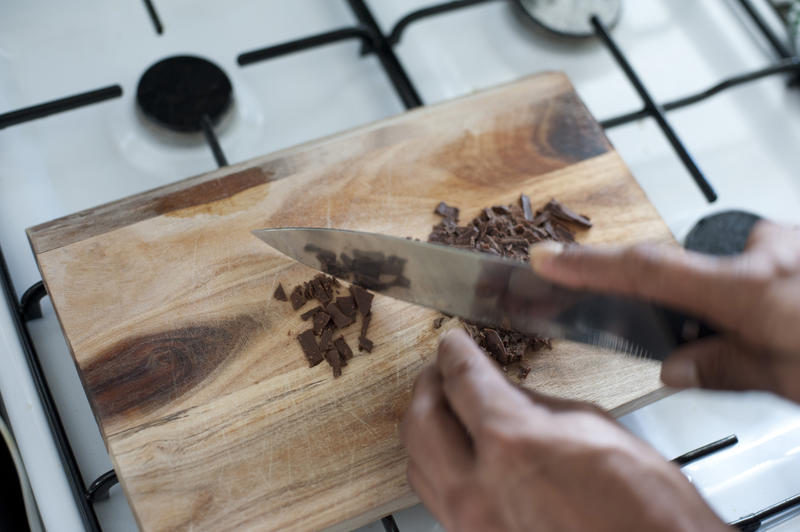 Cooking with chocolate as a cook uses a large sharp knife to chop a bar of chocolate candy on a wooden cutting board