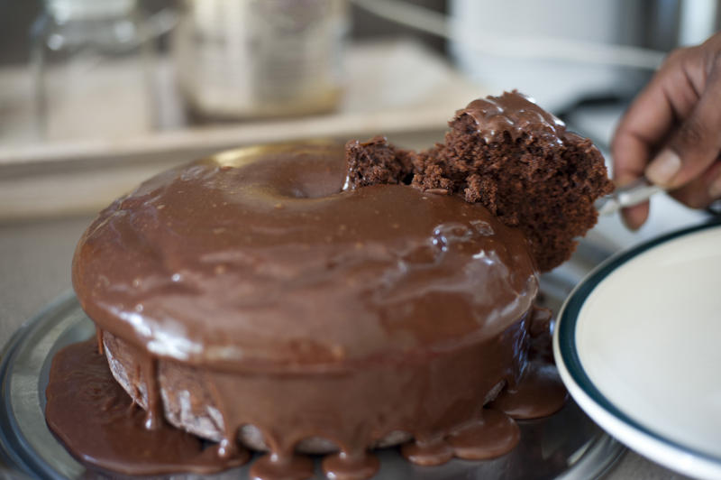 Person serving a freshly baked chocolate sponge cake with oodles of icing dripping onto the plate removing the first slice with a knife