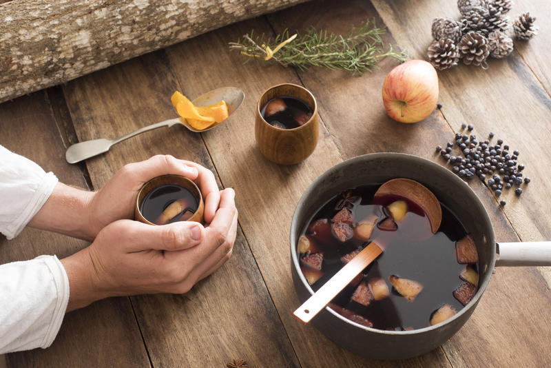 Person warming their hands on a mug of homemade mulled red wine with fresh fruit, herbs and spices surrounding a pot of the spicy aromatic beverage