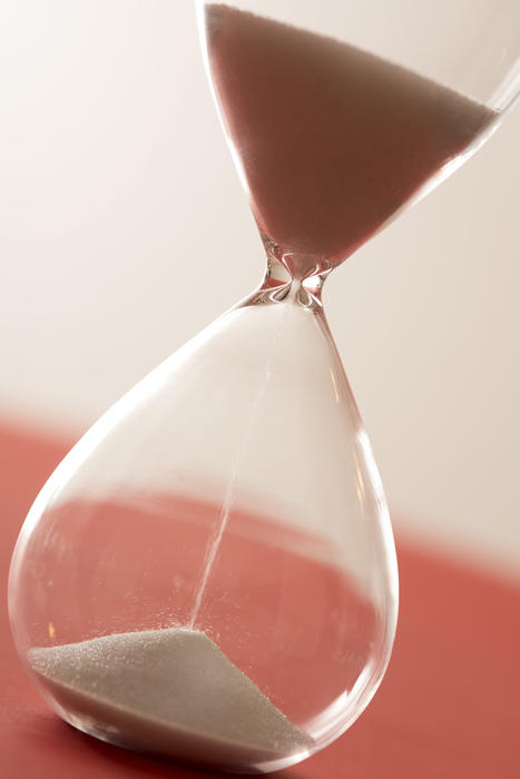 Close up cropped angled view on large glass hourglass with sand pouring close up over red surface