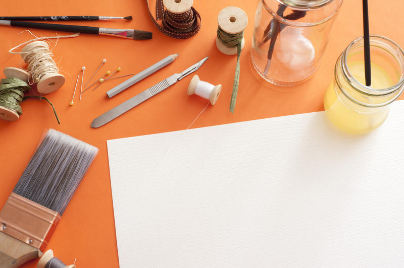 Paintbrushes, pins, wire and other assorted painting tools around blank white sheet of paper with copy space over orange background