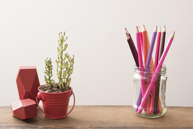 Desk still life with red ornaments and a potted plant in a little bucket alongside a jar of colored pencils against a white wall background