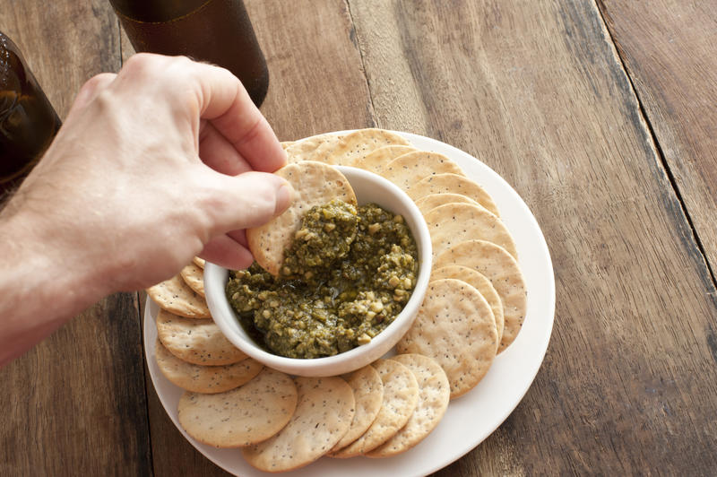First person perspective of hand with cracker plunging into pesto dip in white bowl on plate against a rustic background