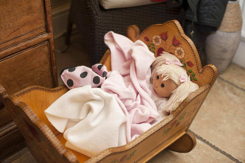 High Angle View of Doll with Blond Hair Nestled Under Pink Blankets in Handmade Wooden Cradle in Family Home