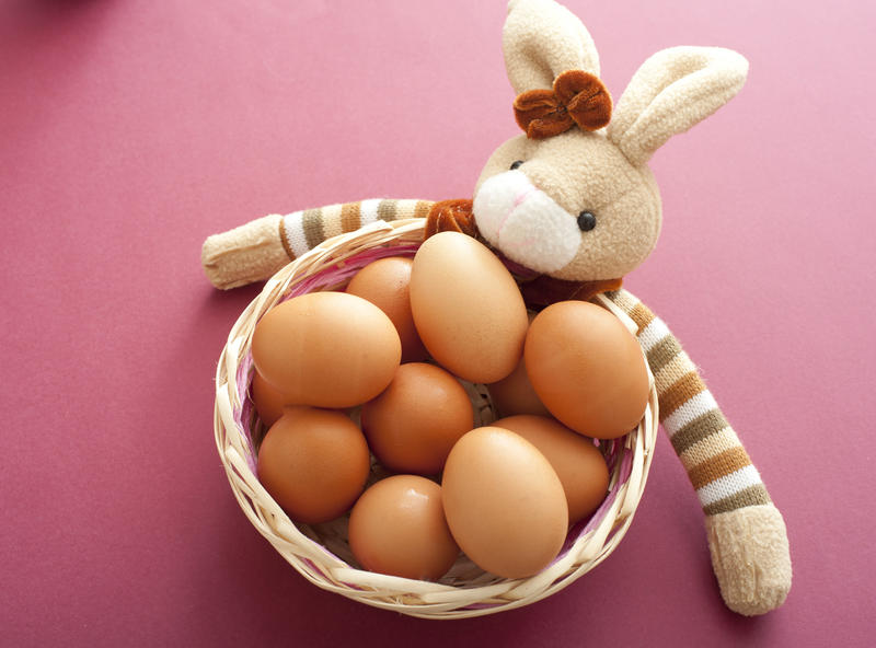 Cute toy Easter bunny with striped arms around a wicker basket filled with fresh brown eggs over a red background