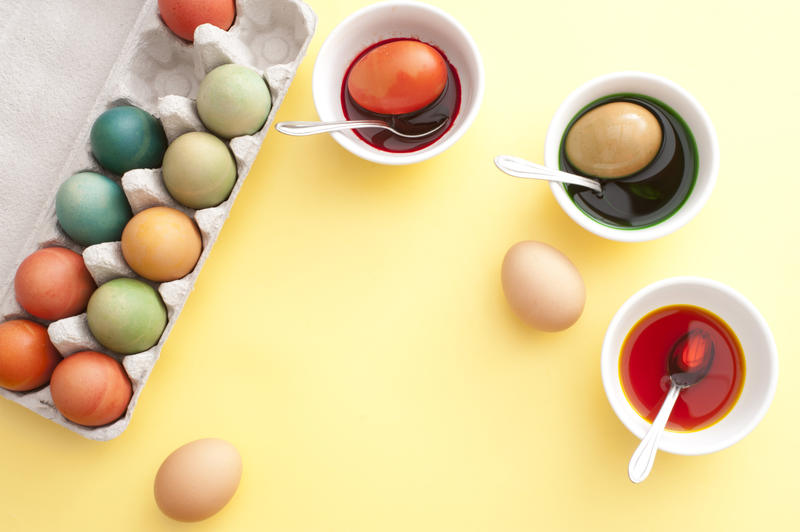 Process of dyeing Easter eggs in bowls of liquid paint with spoons, carton box of colored eggs over yellow table background with copy space in center