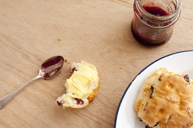 First person perspective on half eaten buttered crispy scone biscuits, spoon, plate and jam in plate over light brown wooden table