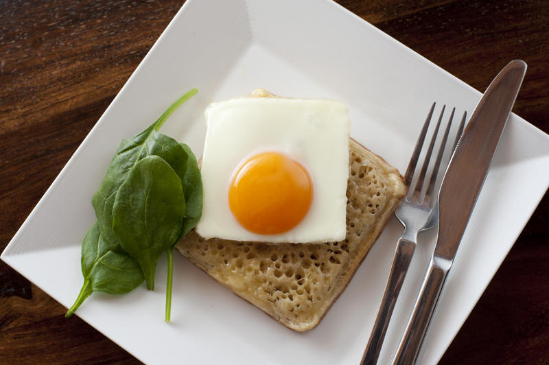 First person perspective view on scrumptious square crumpet with fried egg beside baby spinach leaves in plate next to fork and knife