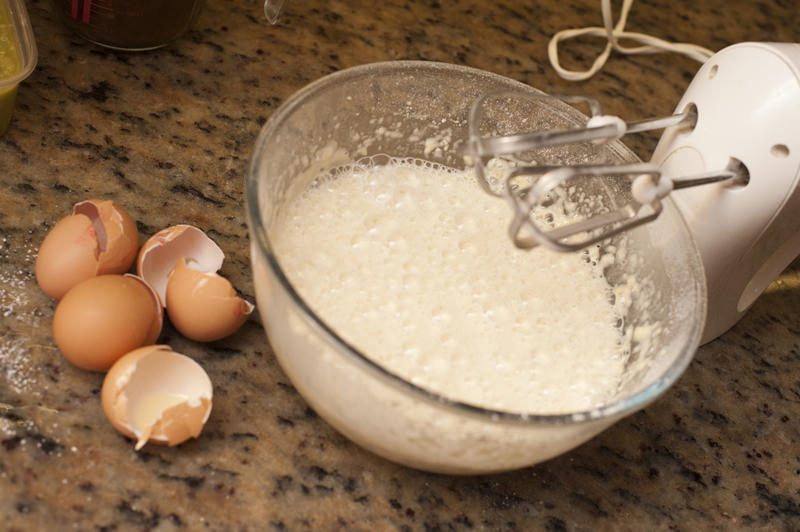 Glass bowl with frothy mixture beside egg shells and electric mixer propped against one side